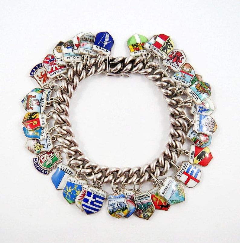 May include: A silver charm bracelet with numerous enameled shield-shaped charms. The charms feature various city names and designs, including "KOLN", "ATHEN", and "COVENTRY". The bracelet is on a white background.