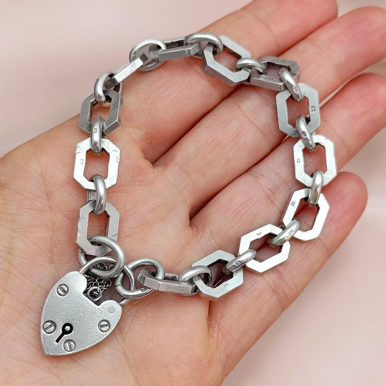 May include: A silver-toned bracelet featuring a heart-shaped padlock clasp and a chain of geometric links. The bracelet is held in a hand against a soft pink background. The padlock has a keyhole and decorative details.