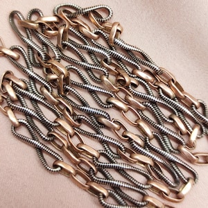 Puede incluir: Primer plano de un collar vintage con eslabones dorados y plateados alternados. La cadena está dispuesta en un patrón en capas sobre un fondo melocotón suave, mostrando su intrincado diseño.