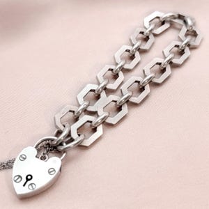 May include: Silver-toned bracelet featuring a heart-shaped padlock charm and a geometric chain design. The padlock has a keyhole and decorative rivets. The chain links are hexagonal. The bracelet is displayed on a light pink surface.