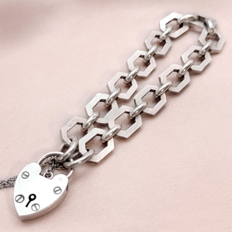 May include: Silver-toned bracelet featuring a heart-shaped padlock charm and a geometric chain design. The padlock has a keyhole and decorative rivets. The chain links are hexagonal. The bracelet is displayed on a light pink surface.