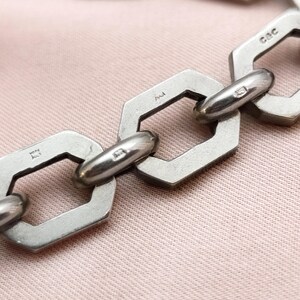 May include: Close-up of a silver chain bracelet with geometric links. The links are hexagonal with rounded connecting bars. The bracelet is on a pink surface. The chain has markings on the links.