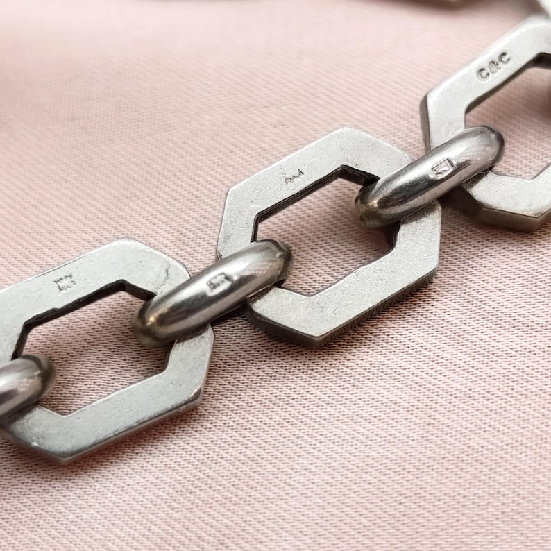 May include: Close-up of a silver chain bracelet with geometric links. The links are hexagonal with rounded connecting bars. The bracelet is on a pink surface. The chain has markings on the links.