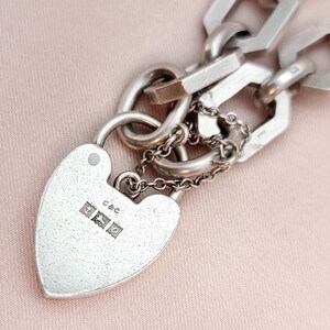 May include: A silver heart-shaped padlock with a small chain and a larger chain link. The padlock has the letters "c&c" and three small hallmarks. The background is a light pink color.