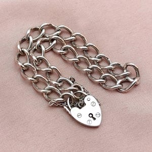 May include: A silver chain necklace with a padlock charm. The chain is made of interlocking links and the padlock has a heart shape and a keyhole.