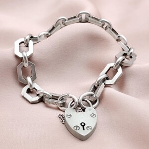 May include: Silver-toned bracelet featuring a heart-shaped padlock clasp. The bracelet has a chain-link design with geometric and circular links. The padlock has a keyhole and decorative rivets. The jewelry is displayed on a pink fabric.