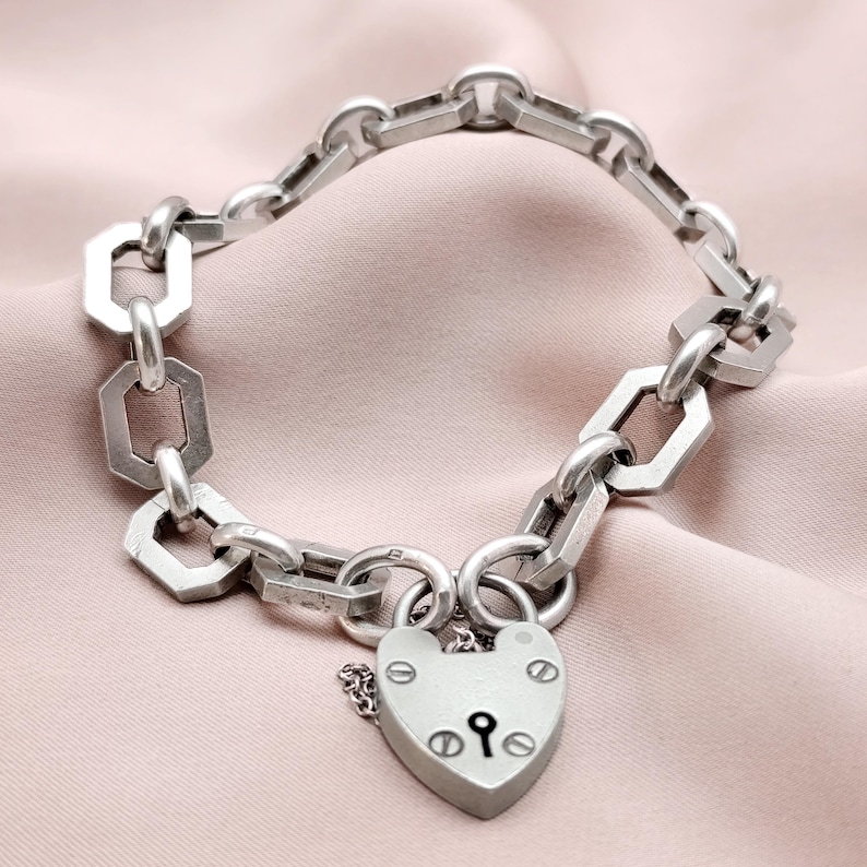 May include: Silver-toned bracelet featuring a heart-shaped padlock clasp. The bracelet has a chain-link design with geometric and circular links. The padlock has a keyhole and decorative rivets. The jewelry is displayed on a pink fabric.
