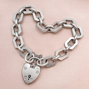 May include: Silver-toned chain bracelet with a heart-shaped padlock charm. The bracelet features geometric links and a keyhole detail on the padlock. The bracelet is displayed on a pink surface.