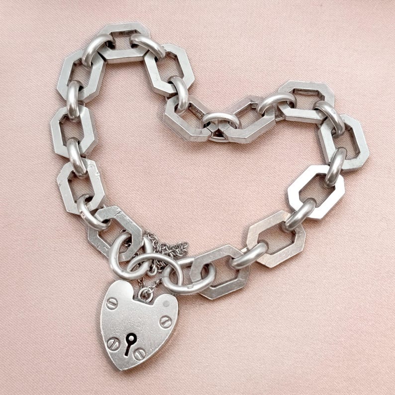 May include: Silver-toned chain bracelet with a heart-shaped padlock charm. The bracelet features geometric links and a keyhole detail on the padlock. The bracelet is displayed on a pink surface.