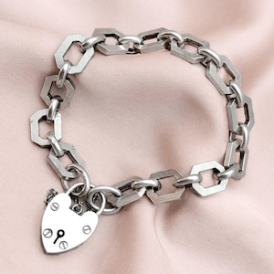 May include: A silver-toned bracelet with a series of geometric links and a heart-shaped padlock charm. The bracelet is displayed on a pink surface, showcasing its intricate design and metallic sheen. The padlock features a keyhole and decorative rivets.