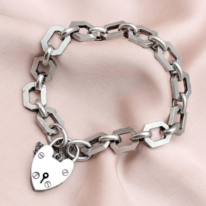 May include: A silver-toned bracelet with a series of geometric links and a heart-shaped padlock charm. The bracelet is displayed on a pink surface, showcasing its intricate design and metallic sheen. The padlock features a keyhole and decorative rivets.