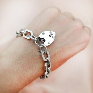 May include: A silver-toned bracelet featuring a heart-shaped padlock charm and a chain-link design. The padlock has a keyhole detail. The bracelet is worn on a wrist, showcasing the jewelry's design and craftsmanship.