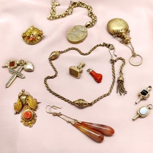 May include: An assortment of antique jewelry pieces, including necklaces, brooches, earrings, and pendants. The collection features gold tones, red coral, and gemstones. The items are arranged on a pink surface.