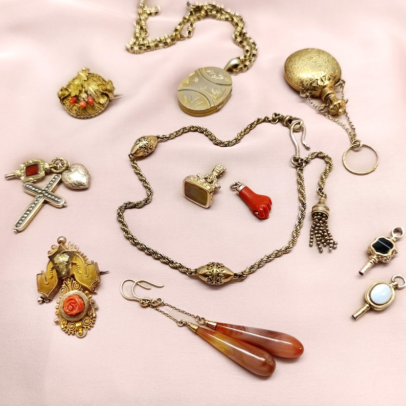 May include: An assortment of antique jewelry pieces, including necklaces, brooches, earrings, and pendants. The collection features gold tones, red coral, and gemstones. The items are arranged on a pink surface.