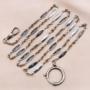 Puede incluir: Cadena de reloj de bolsillo plateada con un diseño decorativo. La cadena presenta eslabones ovalados alargados con detalles en forma de diamante, conectados por eslabones de cadena más pequeños. Un cierre de resorte y un guardapelo circular completan el diseño.