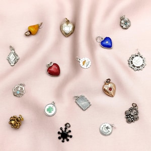 May include: A collection of vintage charms in various shapes and designs, including hearts, flowers, and geometric forms. The charms are made of gold, silver, and other materials, with some featuring colorful enamel and gemstones. The charms are displayed on a pink fabric.