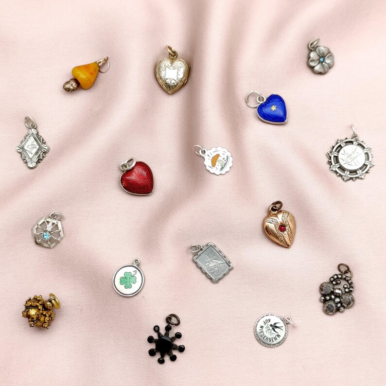 May include: A collection of vintage charms in various shapes and designs, including hearts, flowers, and geometric forms. The charms are made of gold, silver, and other materials, with some featuring colorful enamel and gemstones. The charms are displayed on a pink fabric.