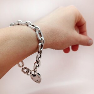 May include: A silver-colored chain bracelet with a heart-shaped padlock charm. The bracelet features a series of rectangular and oval links. The padlock charm has engraved details. The bracelet is worn on a wrist.