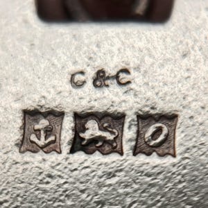 May include: Close-up of a silver object with three hallmarks and the letters "C & C". The hallmarks include an anchor, a lion, and a letter "O" inside rectangular frames. The surface has a textured appearance.