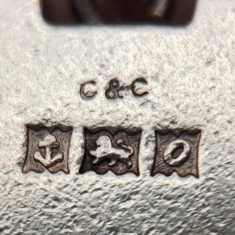 May include: Close-up of a silver object with three hallmarks and the letters "C & C". The hallmarks include an anchor, a lion, and a letter "O" inside rectangular frames. The surface has a textured appearance.
