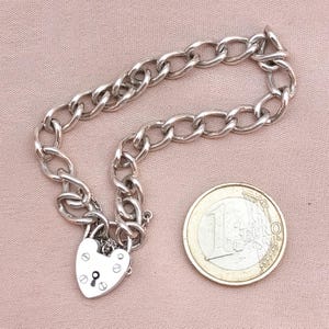 May include: A silver chain bracelet with a heart-shaped padlock charm. The bracelet is about the size of a Euro coin.