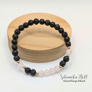 Könnte beinhalten: Armband aus schwarzen Lavasteinen und rosafarbenem Quarz mit goldenen Akzenten. Das Armband liegt auf einer Holzoberfläche. "Schmuck'n Roll Soul und Energie Schmuck" ist auf der Oberfläche geschrieben.