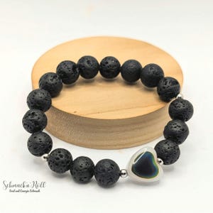 Könnte beinhalten: Schwarzes Lava-Stein-Perlenarmband mit herzförmigem Anhänger. Das Armband hat runde, poröse schwarze Perlen und einen herzförmigen Anhänger mit schillernden Farben. Das Armband wird auf einer Holzoberfläche präsentiert. Der Text "Schmuck'n'Roll" ist sichtbar.