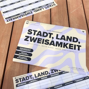 K&ouml;nnte beinhalten: Mehrere bedruckte Papierb&ouml;gen mit dem Text "STADT, LAND, ZWEISAMKEIT" in gro&szlig;en schwarzen Buchstaben. Die B&ouml;gen haben ein hellviolettes und cremefarbenes Wellendesign. Die B&ouml;gen liegen auf einer Holzoberfl&auml;che.