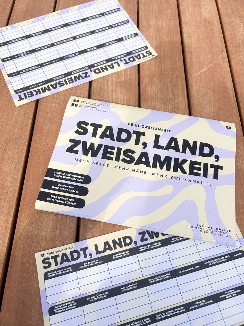 K&ouml;nnte beinhalten: Mehrere bedruckte Papierb&ouml;gen mit dem Text "STADT, LAND, ZWEISAMKEIT" in gro&szlig;en schwarzen Buchstaben. Die B&ouml;gen haben ein hellviolettes und cremefarbenes Wellendesign. Die B&ouml;gen liegen auf einer Holzoberfl&auml;che.