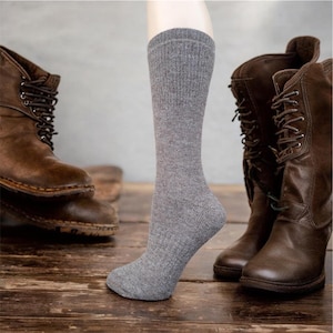 May include: A pair of gray crew socks with a ribbed texture. The socks are shown on a model's leg with brown leather boots in the background.