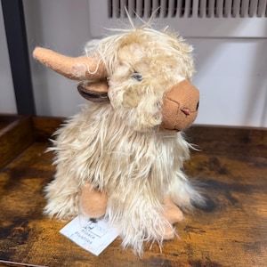 May include: A plush Highland cow toy with shaggy, light brown fur. It has small, curved horns, a brown snout, and small, dark eyes. A tag that reads "Alpaca Plushies" is attached to the toy.