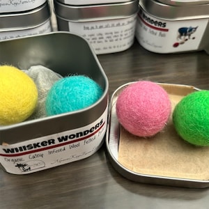 May include: A set of four felt catnip balls in a silver tin. The balls are yellow, turquoise, pink, and green. The tin has a label that reads "Whisker Wonders Organic Catnip Infused Wool Felted Balls Alpaca Key Farm Co."