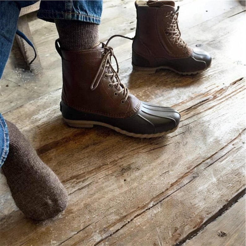 May include: A pair of brown leather lace-up boots with a black rubber bottom. The boots are worn by a person standing on a wooden floor.