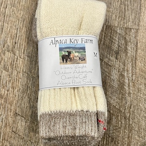 May include: Cream-colored alpaca boot socks, folded and banded with a label. The label reads "Alpaca Key Farm" and "Heavy-Weight Outdoor Adventure Over-the-Calf Alpaca Boot Sock." The socks have a brown and cream-colored cuff and a red accent.