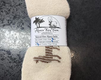 May include: A pair of cream-colored natural fiber alpaca socks with a brown alpaca graphic. The socks are wrapped in a white paper label featuring the text Alpaca Key Farm, Natural Fiber Alpaca Socks, and checkboxes for style, weight, and size.