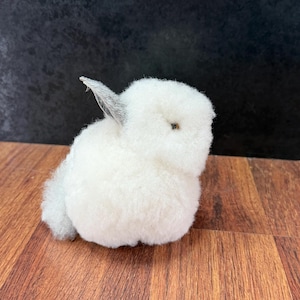 May include: A small, fluffy white rabbit figurine with a rounded body, soft textured fur, a single visible pointed ear with grey detailing, and a small dark eye. The rabbit is positioned in a seated posture, showcasing a dense, wool-like appearance.