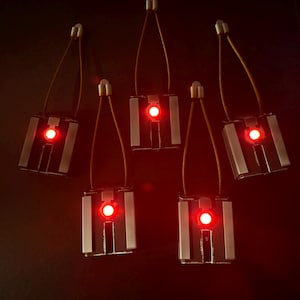 May include: Five rectangular metallic pendants with a central glowing red circular light, each attached to a thin brown cord loop. The pendants feature a silver-toned finish with vertical detailing and a small metallic clasp at the top of the cord.