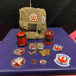 May include: An assortment of Star Wars themed merchandise, including a canvas backpack, two red and black canisters, patches, and pins. The patches feature various logos and text, and the items are arranged on a purple surface.