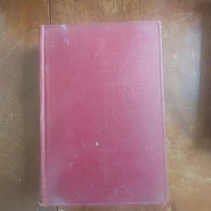 以下が含まれることがあります： 色あせた赤い表紙の長方形のハードカバー本。本の表紙はわずかにテクスチャ加工されており、小さな白い斑点など、摩耗の兆候が見られます。背表紙は左側にあり、本の製本を示しています。