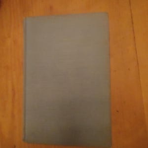 Puede incluir: Un libro cerrado con una cubierta gris, apoyado sobre una superficie de madera. La cubierta del libro es de un color gris sólido y apagado, y los bordes están ligeramente desgastados. La superficie de madera tiene un tono marrón-anaranjado cálido.