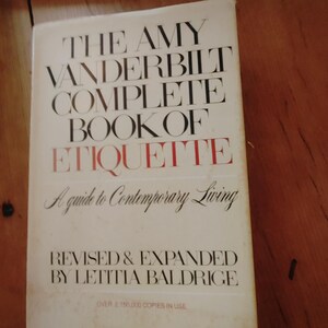 May include: A vintage hardcover book, "THE AMY VANDERBILT COMPLETE BOOK OF ETIQUETTE." The cover is white with black and red lettering. It's a guide to contemporary living, revised and expanded by Letitia Baldrige.
