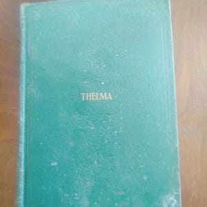 May include: A vintage book with a teal cover. The title "THELMA" is embossed in white, centered on the cover. The cover has a decorative embossed design above the title. The book shows signs of wear and age.