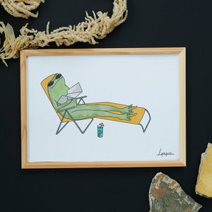 May include: A framed art print featuring a cartoon frog relaxing on a beach chair, wearing sunglasses and reading. The frog is green, the chair is yellow and orange, and a drink with a straw sits nearby. The artwork is set against a white background.
