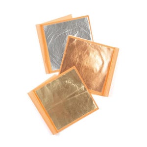 May include: Three square sheets of metallic leaf in silver, copper, and gold tones. Each thin, reflective sheet is mounted on a light orange paper backing, displaying a crinkled, delicate texture across the surface of the metallic material.