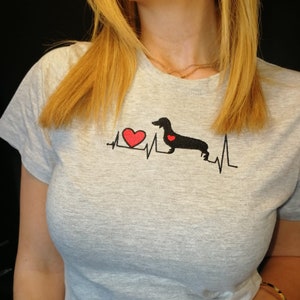 K&ouml;nnte beinhalten: Graues T-Shirt mit einer schwarzen Dackel-Silhouette und einem roten Herzen auf einer Herzschlag-Liniengrafik. Der Text "Ich liebe meinen Dackel" ist auf das T-Shirt gedruckt.