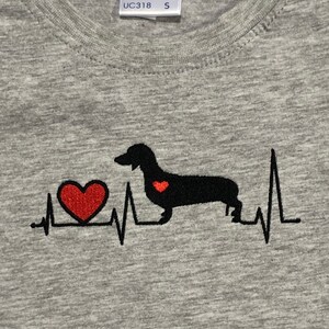 K&ouml;nnte beinhalten: Graues T-Shirt mit einer schwarzen Dackel-Silhouette und einem roten Herzen. Der Dackel hat ein kleines rotes Herz auf der Brust. Das Design ist eine Herzschlaggrafik mit dem Dackel in der Mitte.