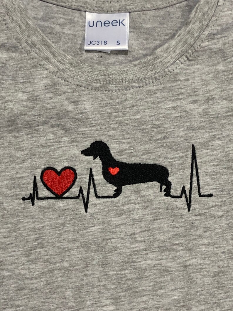 K&ouml;nnte beinhalten: Graues T-Shirt mit einer schwarzen Dackel-Silhouette und einem roten Herzen. Der Dackel hat ein kleines rotes Herz auf der Brust. Das Design ist eine Herzschlaggrafik mit dem Dackel in der Mitte.