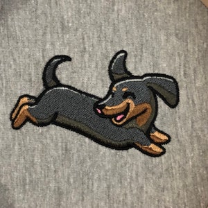 May include: Embroidered patch of a black and brown dachshund running with a happy expression. The patch is on a gray background.