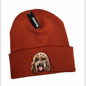 May include: A rust-coloured beanie hat with a folded brim. The front of the hat has an embroidered image of a dog's face with a pink tongue. A black tag with white text that reads "Beechfield ORIGINAL HEADWEAR" is attached.