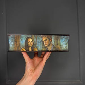Puede incluir: Una caja rectangular con una imagen pintada de dos personas en un bosque. La caja tiene un borde oscuro. La imagen muestra a un hombre y una mujer con un fondo de bosque. La caja está sostenida por una mano.